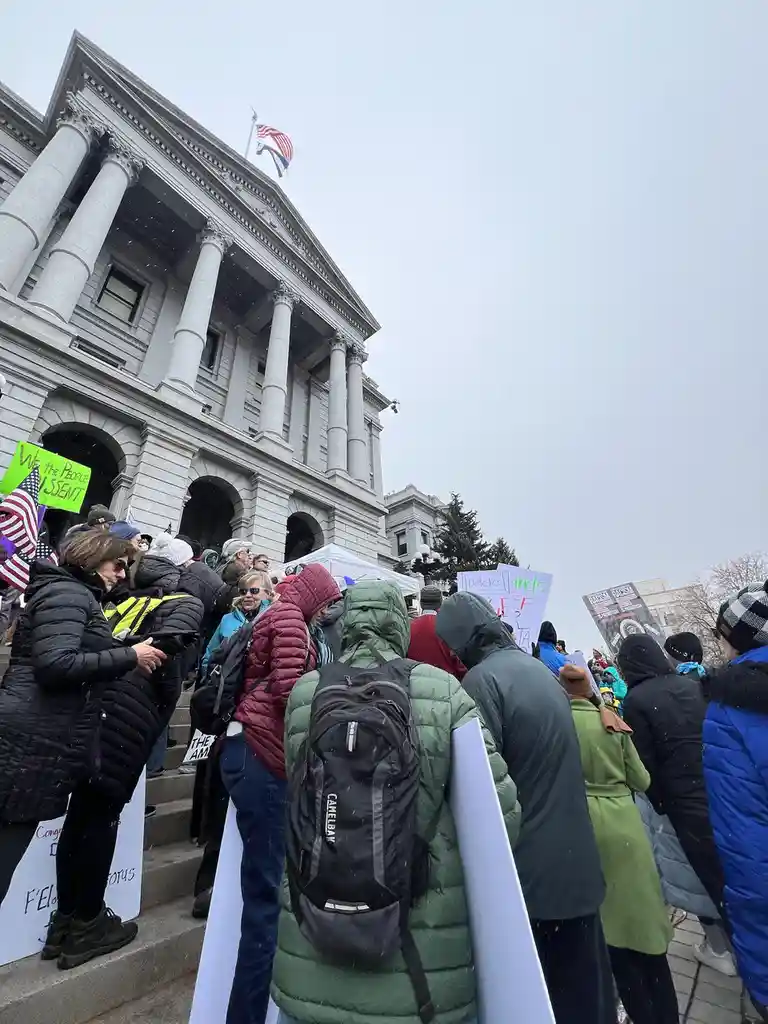 A crowd of protesters in winter coats stands outside a large classical-style government building with columns and an…