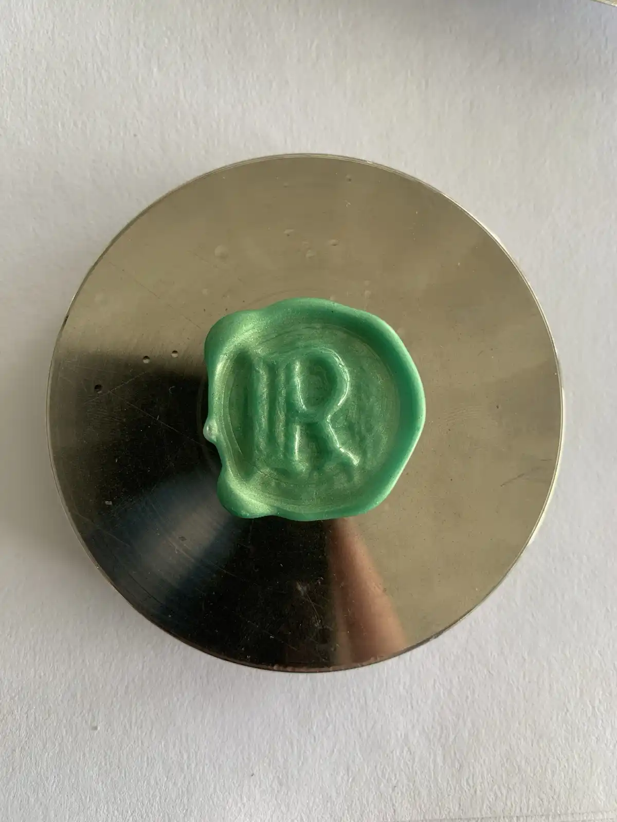 A shiny green wax seal embossed with the letter 'R' sits centered on a reflective gold-colored vinyl record. The record…
