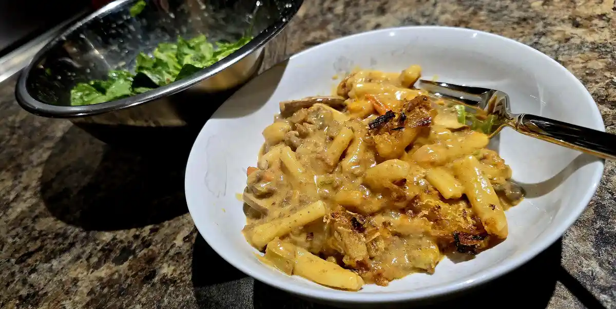 A white bowl of creamy, cheesy pasta with some browned bits sits on a granite countertop, next to a metal bowl of green…