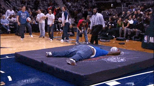 A sequence of six images shows a man in a blue shirt and jeans lying on a large black mat on a basketball court. He then…
