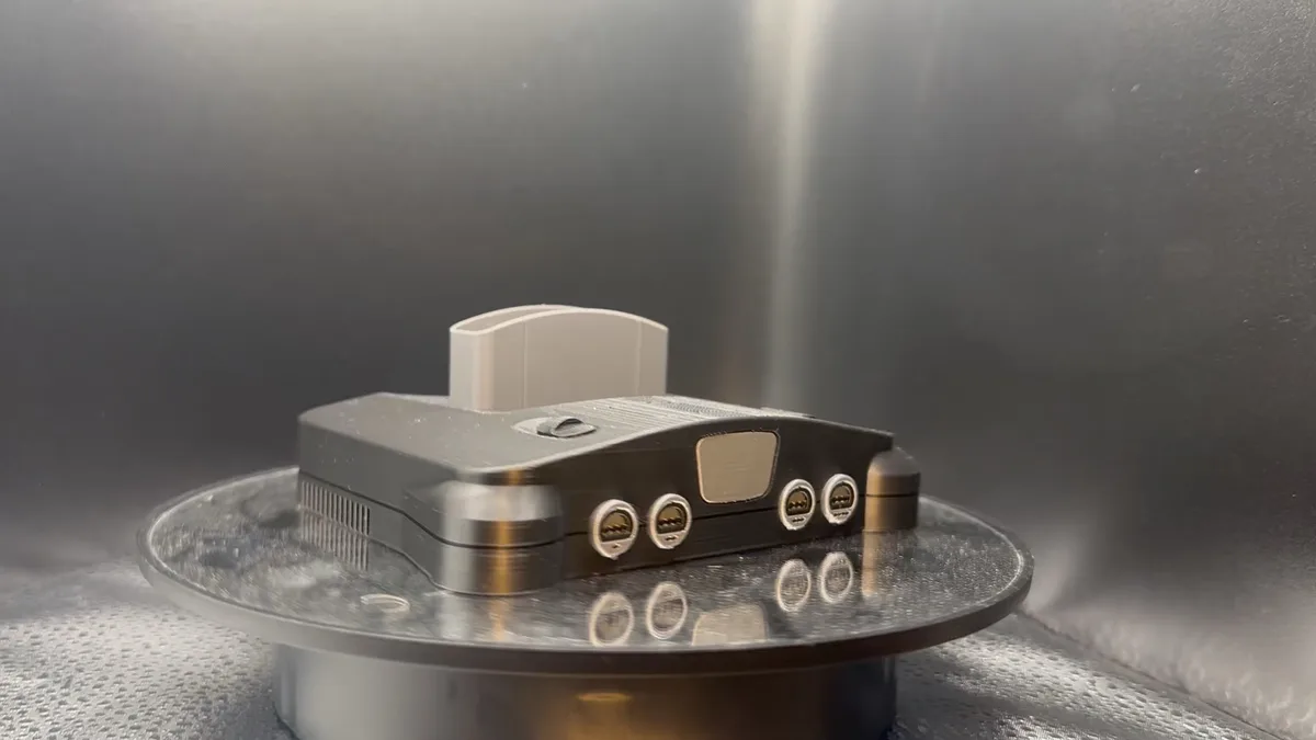 A metallic, 3D-printed replica of a Nintendo 64 console sits on a rotating display stand inside what appears to be a 3D…
