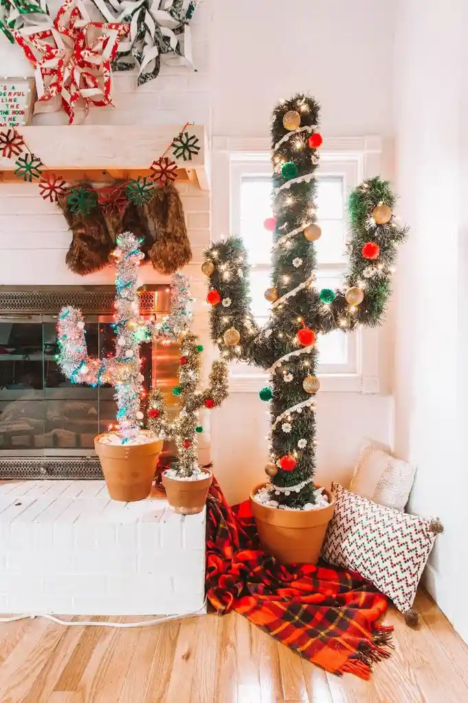 A festive indoor scene features two cacti decorated as Christmas trees, adorned with lights, ornaments, and tinsel, placed…