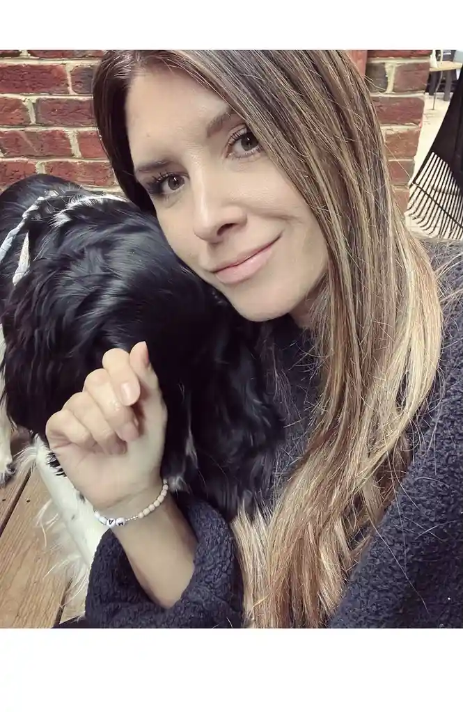 A woman with long brown hair and a dark sweater is smiling gently while holding a black and white dog close to her face. Her…
