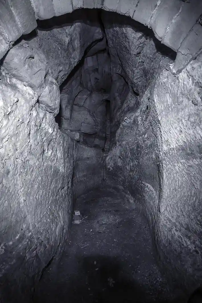 A black-and-white photo shows a narrow, dimly lit stone tunnel with rough, uneven walls and a dark, shadowy path receding…