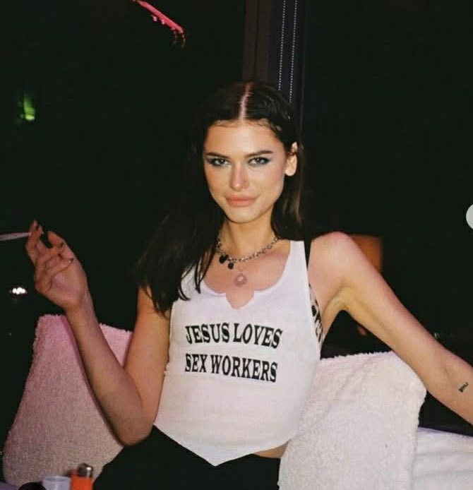 A woman with dark hair and makeup sits on a couch wearing a white crop top with the text 'JESUS LOVES SEX WORKERS' printed…
