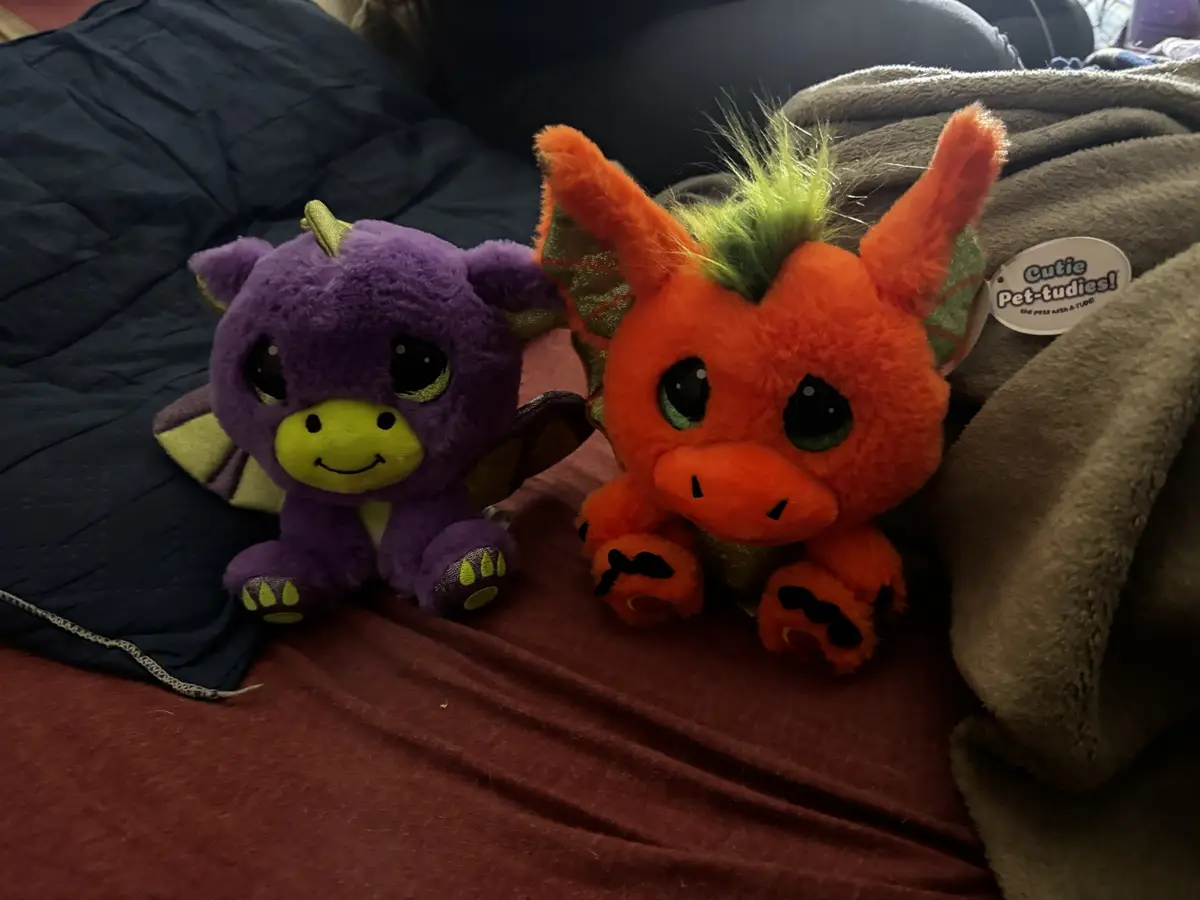 Two small, fluffy stuffed dragons sit side-by-side on a bed with blankets. The dragon on the left is purple with a yellow…