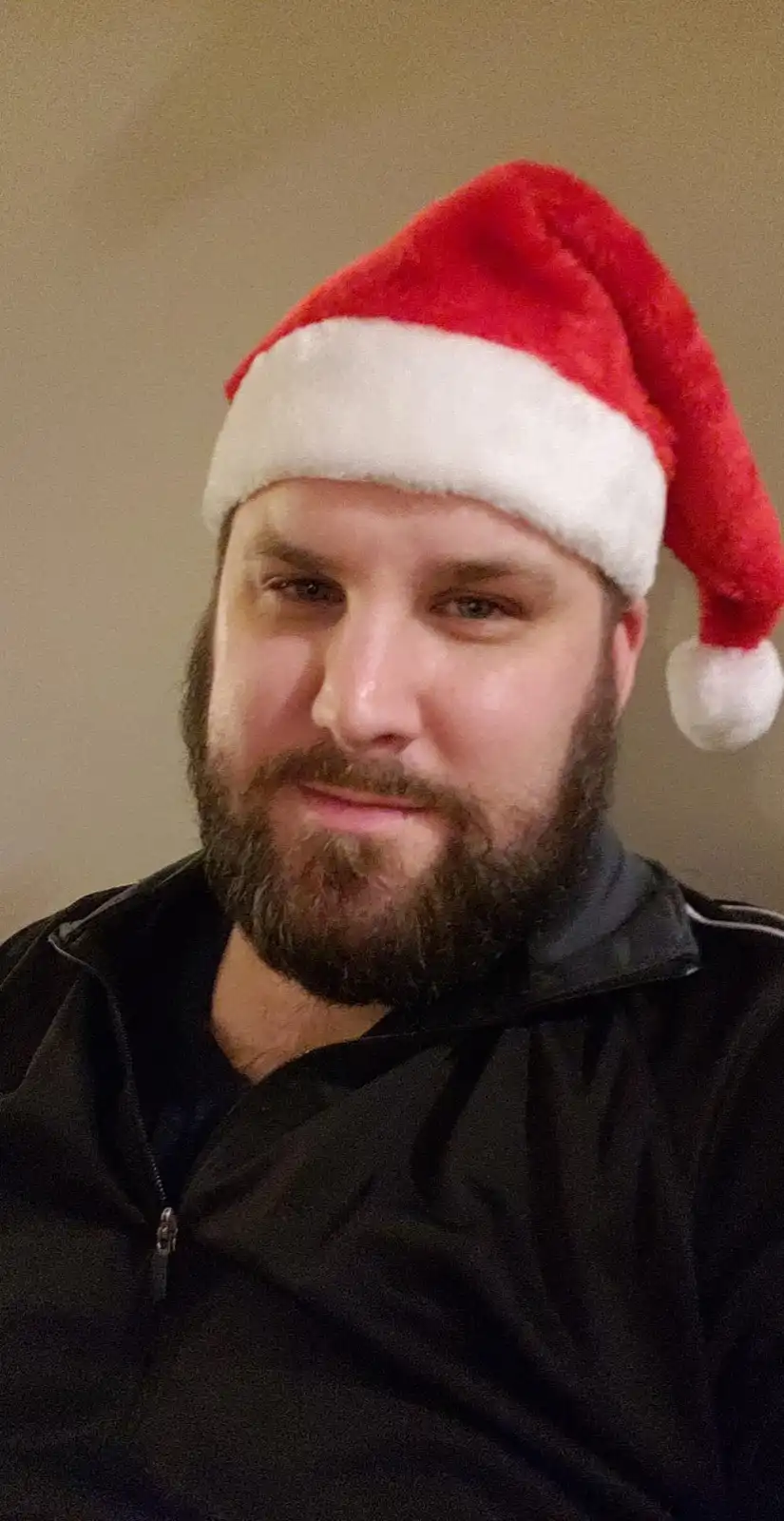 A man with a beard wearing a red and white Santa hat looks at the camera with a subtle, unenthusiastic expression. He is…