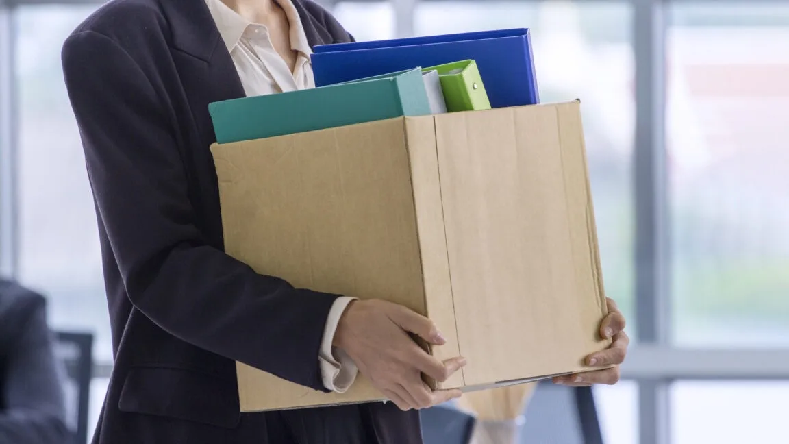 a person in business attire is holding a cardboard box overflowing with colorful binders and folders, suggesting they are…