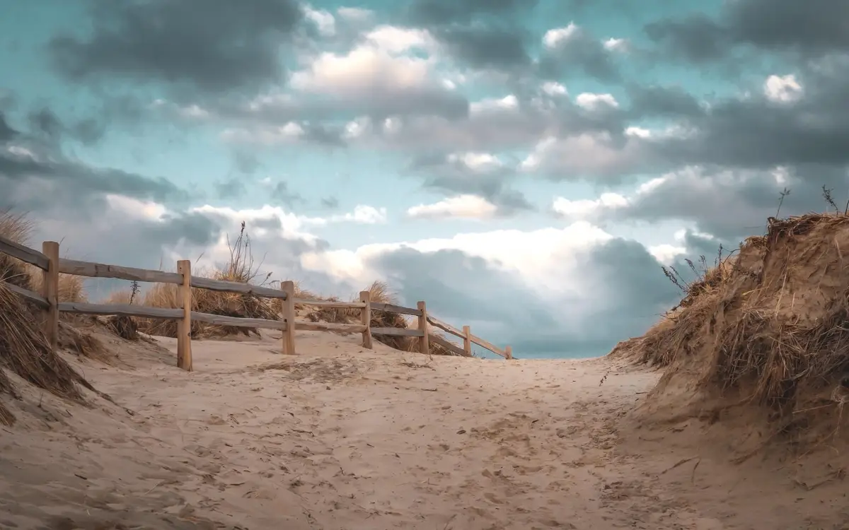 A scenic view of a sandy dune path with a wooden fence on the left, leading toward a cloudy sky. The sand is textured with…