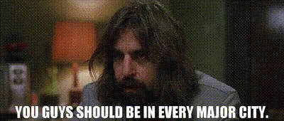 The image shows a man with long brown hair and a beard, wearing a gray shirt, sitting in a dimly lit room with a lamp and…