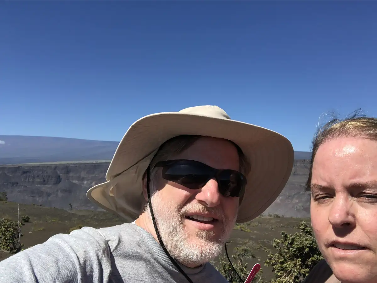A selfie of two people outdoors, one man wearing a wide-brimmed hat and sunglasses smiling at the camera, the other person…