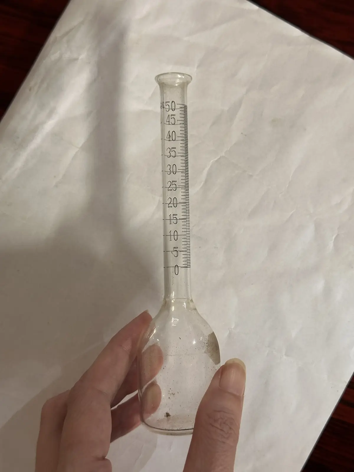 A hand holds a clear glass graduated cylinder with measurement markings from 0 to 50, resting on white paper. The overlaid…