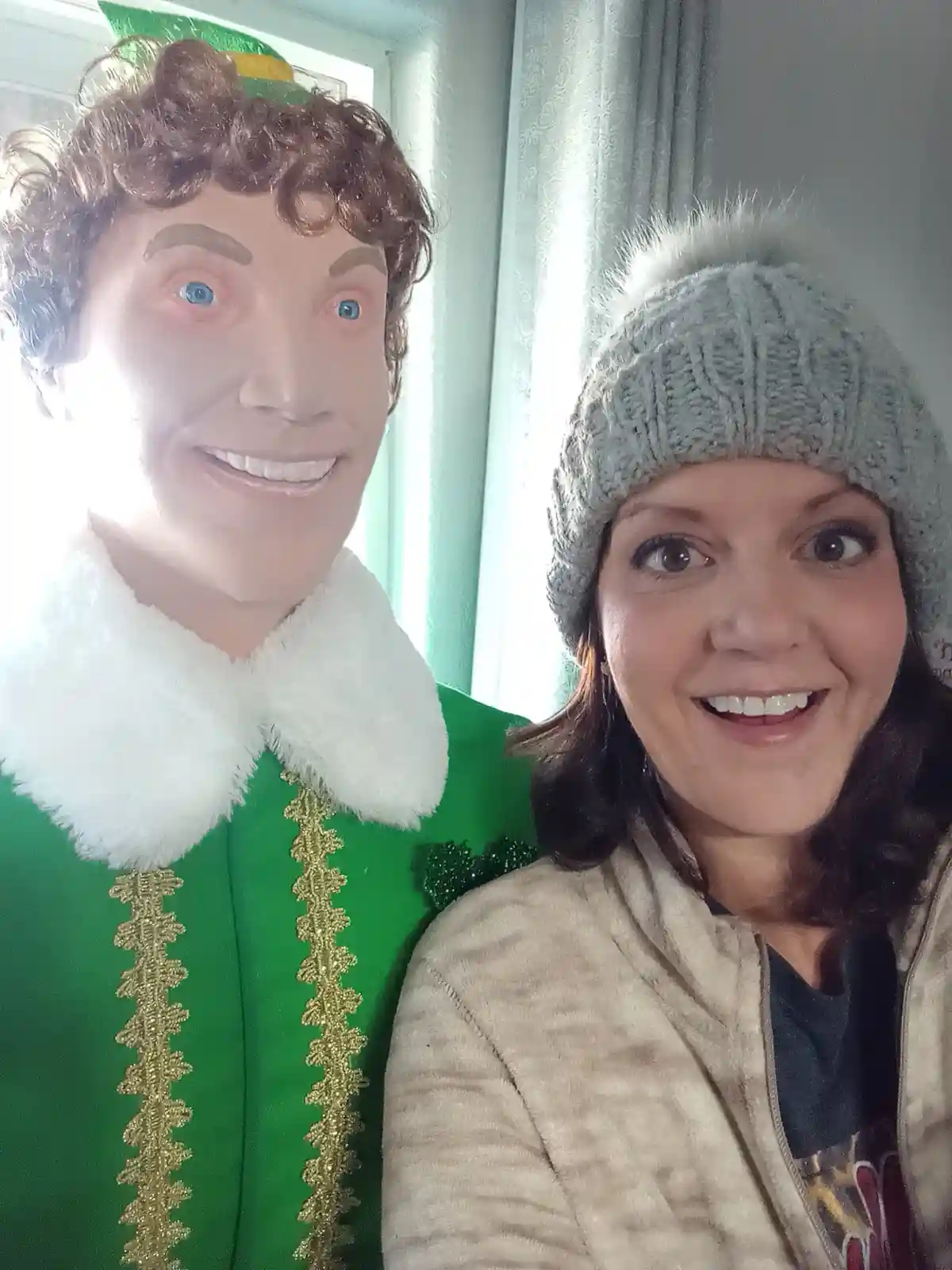 A woman in a grey knit beanie smiles beside a life-sized, smiling Buddy the Elf mannequin in a green suit with white fur…