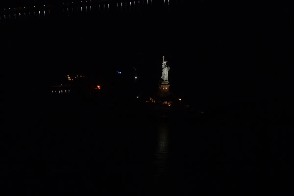 A dark nighttime photo shows the Statue of Liberty illuminated against a black background, with water reflecting distant…