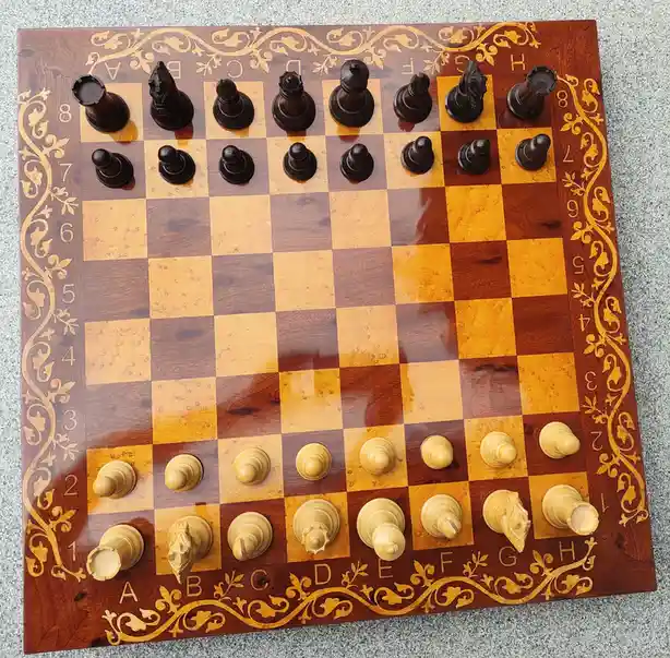 An ornate wooden chessboard with dark and light wood squares, decorated with gold inlaid floral patterns along the edges and…