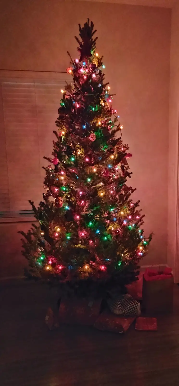 A full, decorated artificial Christmas tree with multicolored lights and ornaments stands in a dimly lit room, surrounded by…