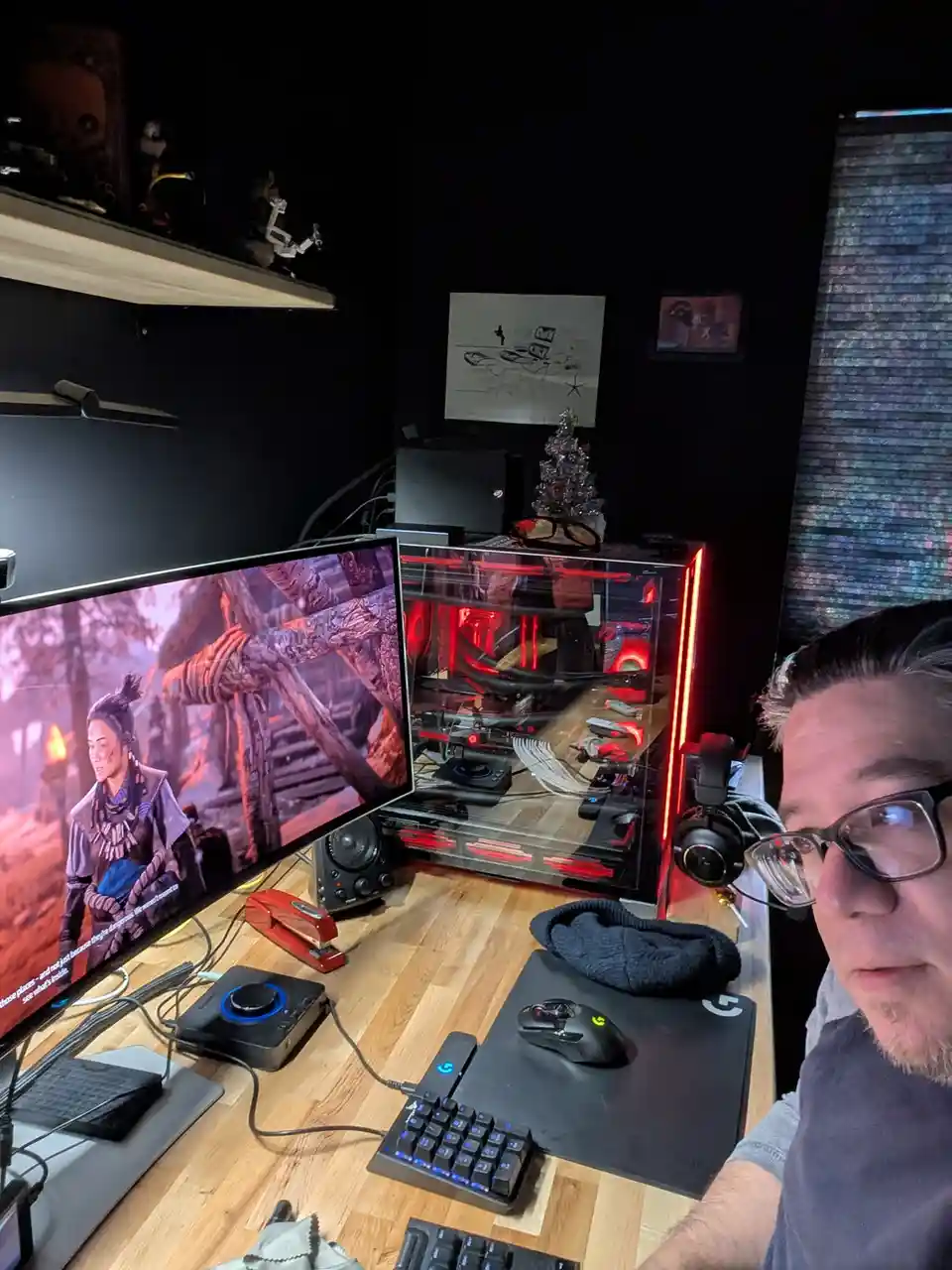 A man with glasses sits at a wooden desk in a dark room, looking at the camera while a large monitor displays a scene from a…
