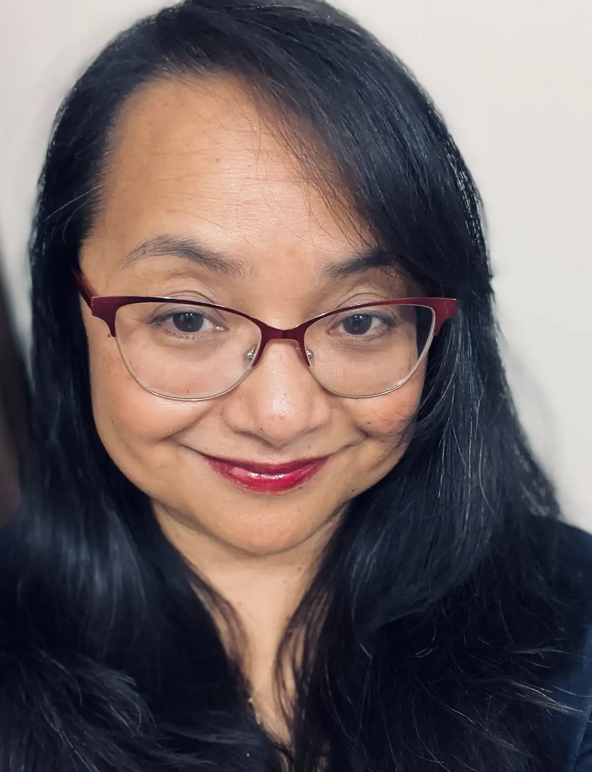 A close-up portrait of a woman with long dark hair, wearing red-framed glasses and a subtle smile, against a plain white…