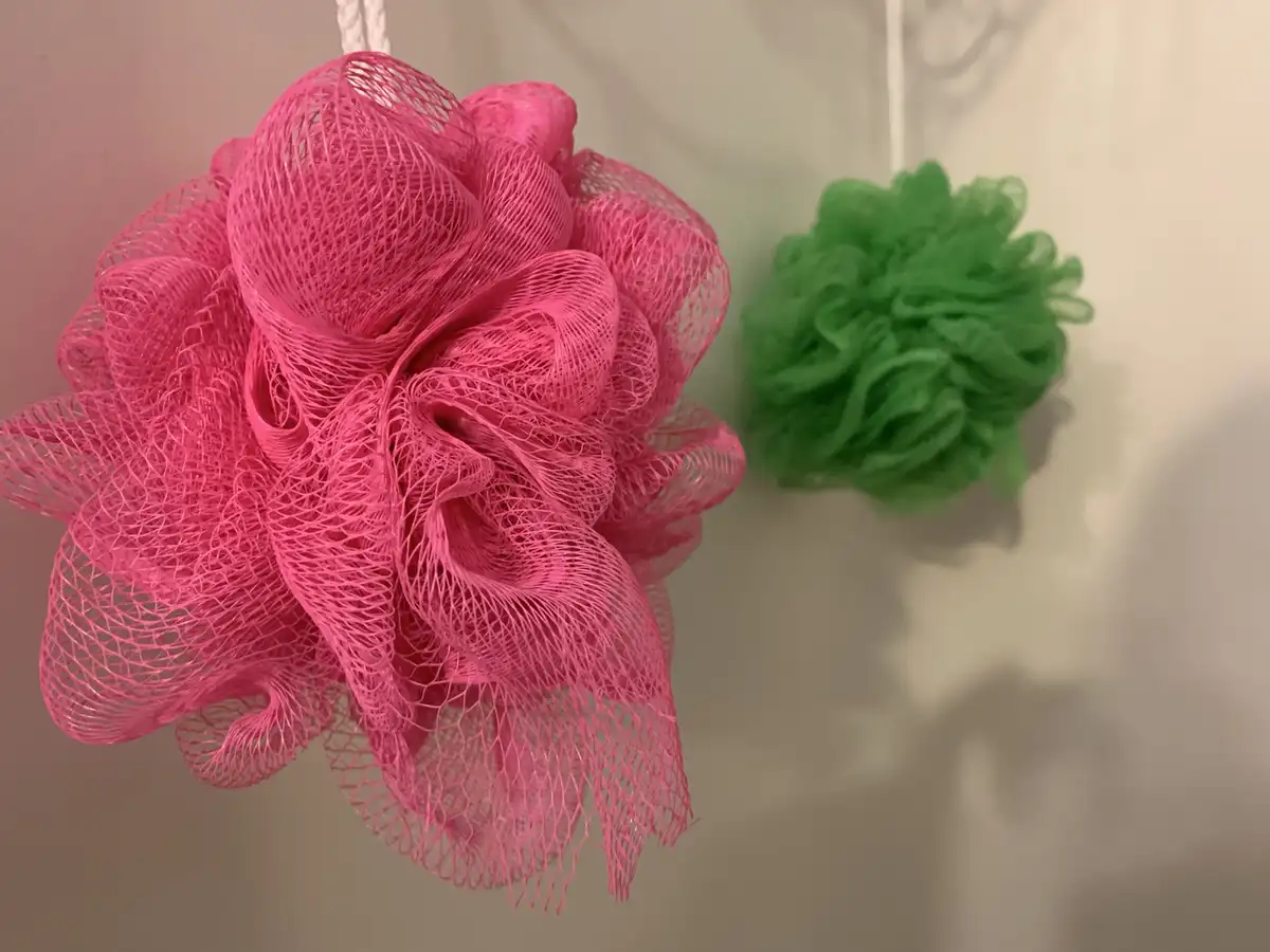 Two bath loofahs hang side by side against a plain wall; a large, elaborate pink loofah dominates the left, while a smaller…