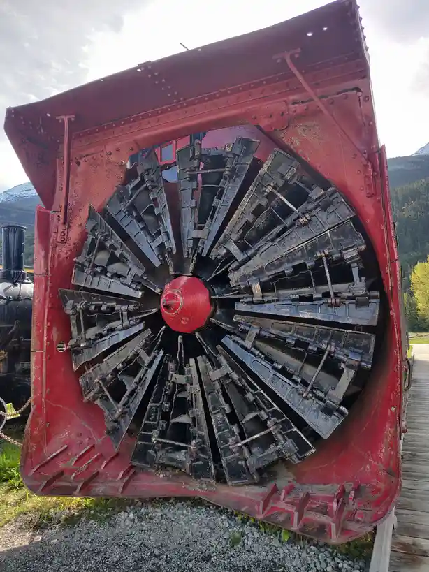A close-up of a large, red, vintage snowplow with a radial blade assembly, showing its heavy-duty construction and weathered…