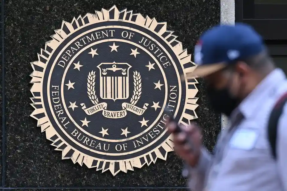 A person in a blue cap and face mask walks past a large, clear FBI seal on a dark granite wall, looking down at their phone…