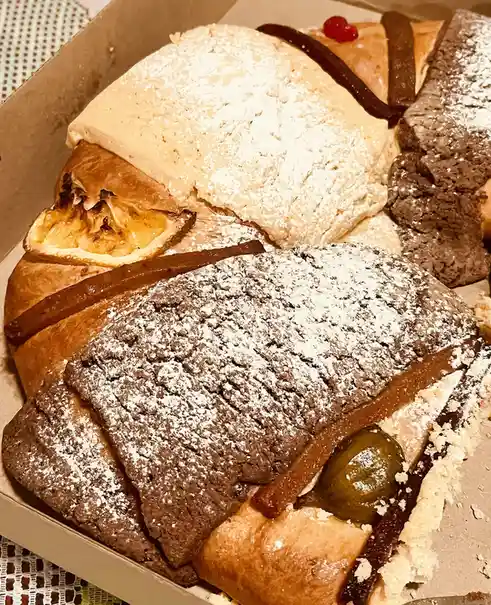 A close-up photo shows a cardboard box filled with various pastries, including chocolate and cream-filled croissants dusted…