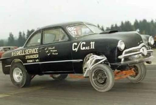 A black vintage race car with 'K-SHELL SERVICE' and 'STU RICKER' written on its side, lifted on large tires with exposed…