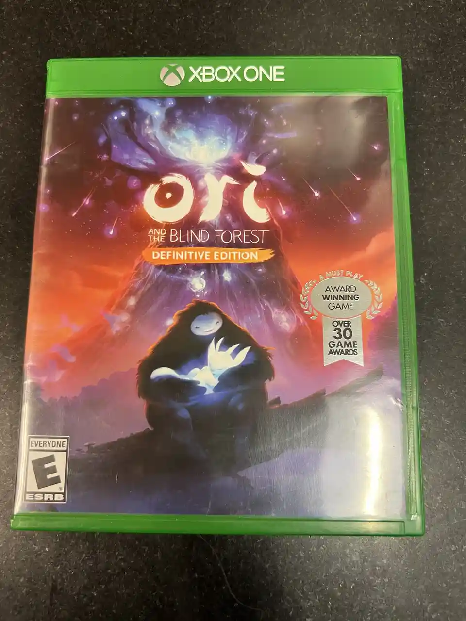 A physical copy of the Xbox One game 'Ori and the Blind Forest: Definitive Edition' is shown in its green case, featuring…