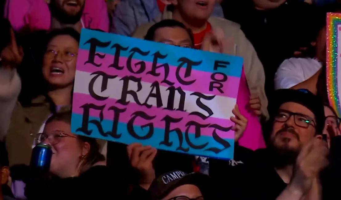 A crowd of people at an event, one person holds up a sign with the text 'FIGHT FOR TRANS RIGHTS' written in black…