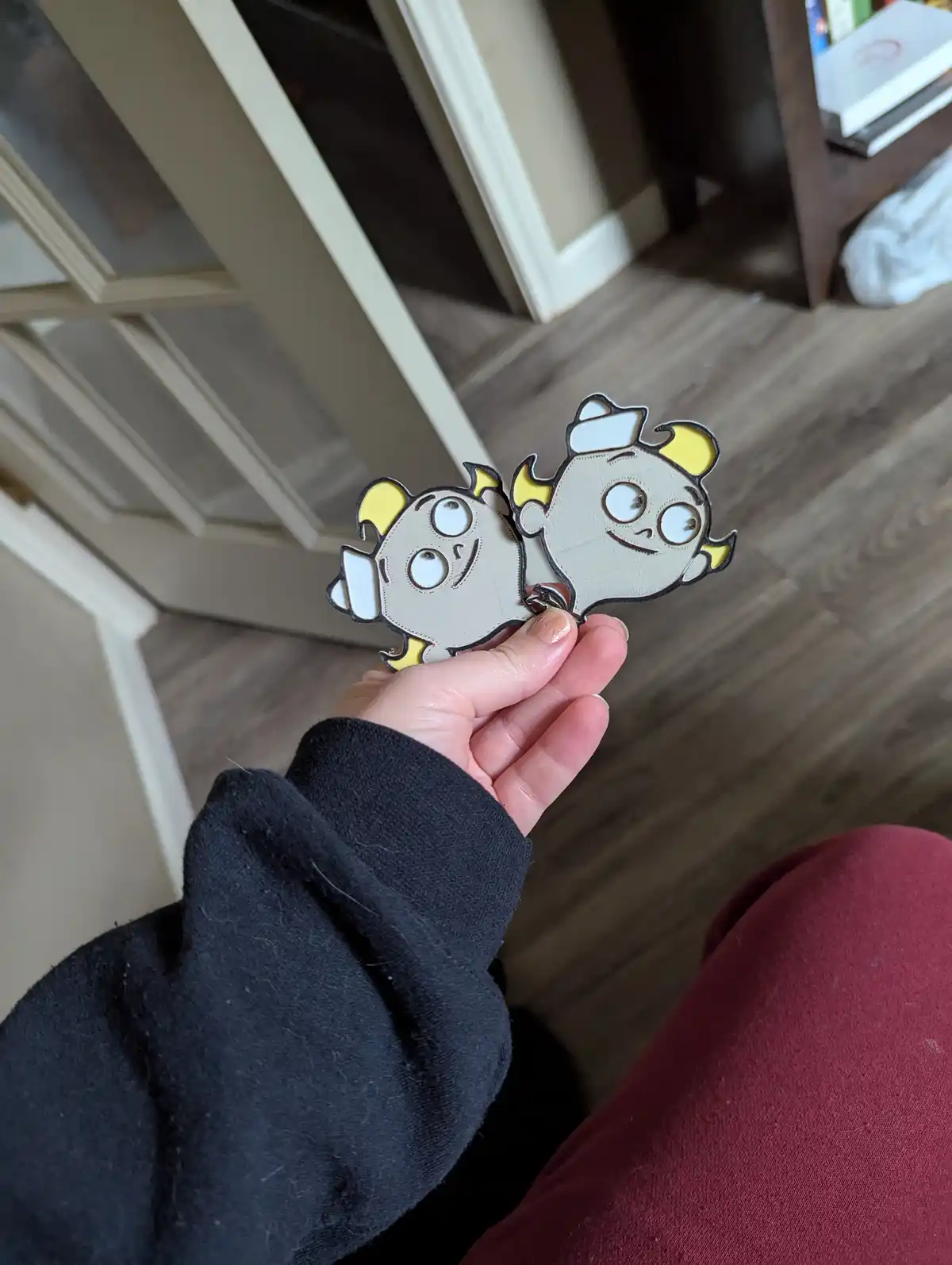 A person holds two identical, cartoonish, gray and yellow character cutouts with large eyes and horns, resembling the…