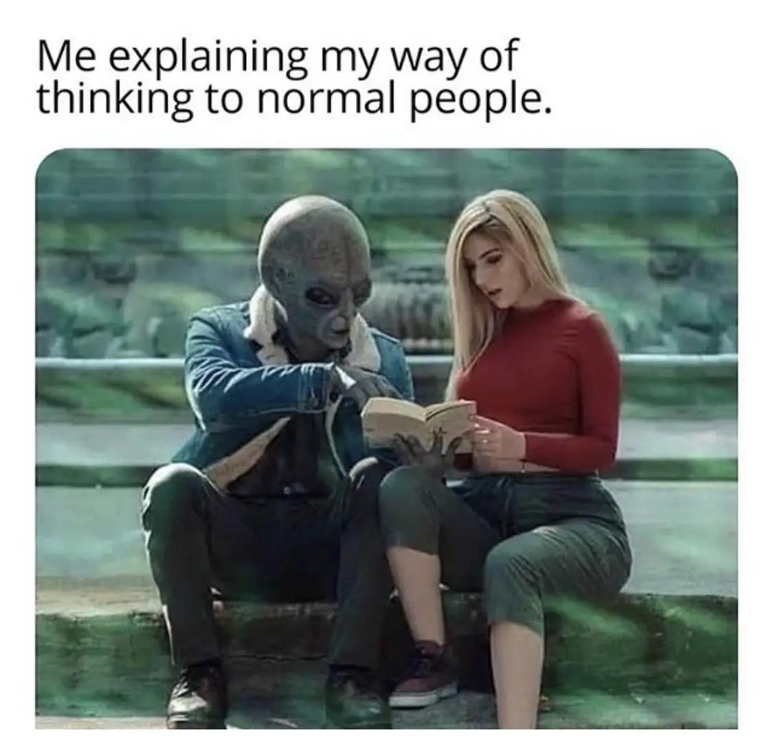 An alien with a large head and grey skin sits next to a human woman in a red shirt, both looking at an open book. The alien…