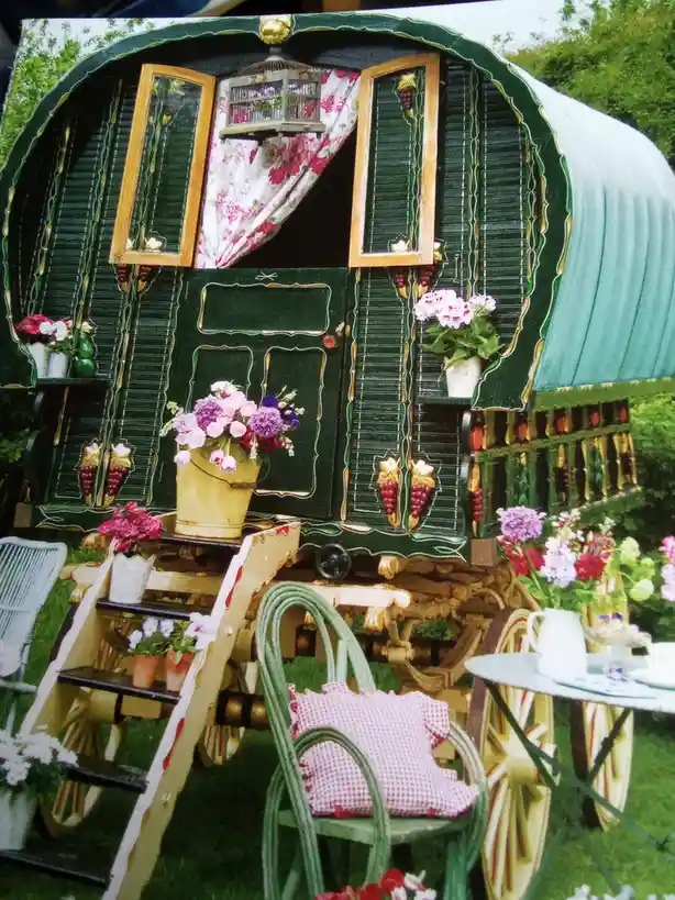 A highly decorated green gypsy caravan with ornate wooden shutters, floral curtains, and a birdcage hanging in the window…