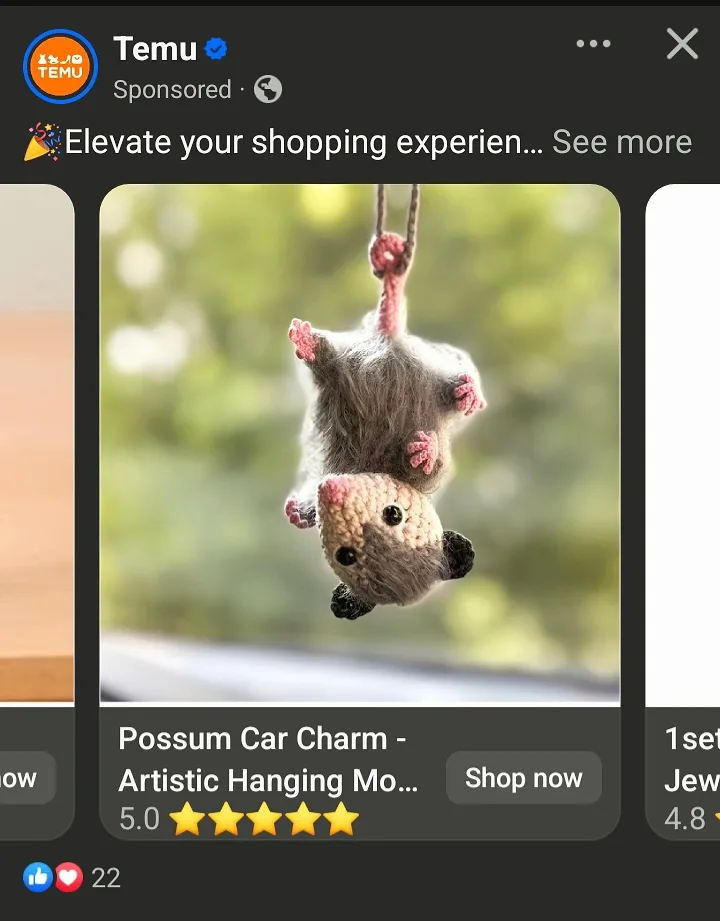 A crocheted possum charm hangs upside down from a string, suspended in front of a blurred green background. The product is…