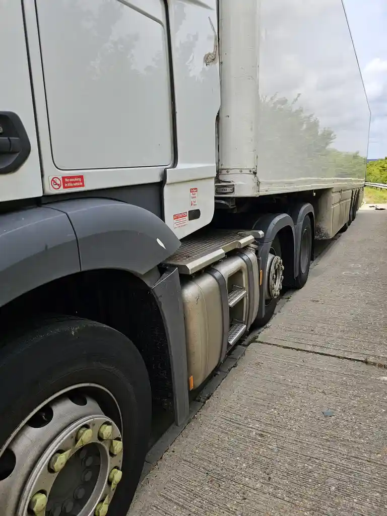 A close-up photo of the side of a large white semi-truck parked next to a concrete shoulder, showing its wheels, fuel tank…