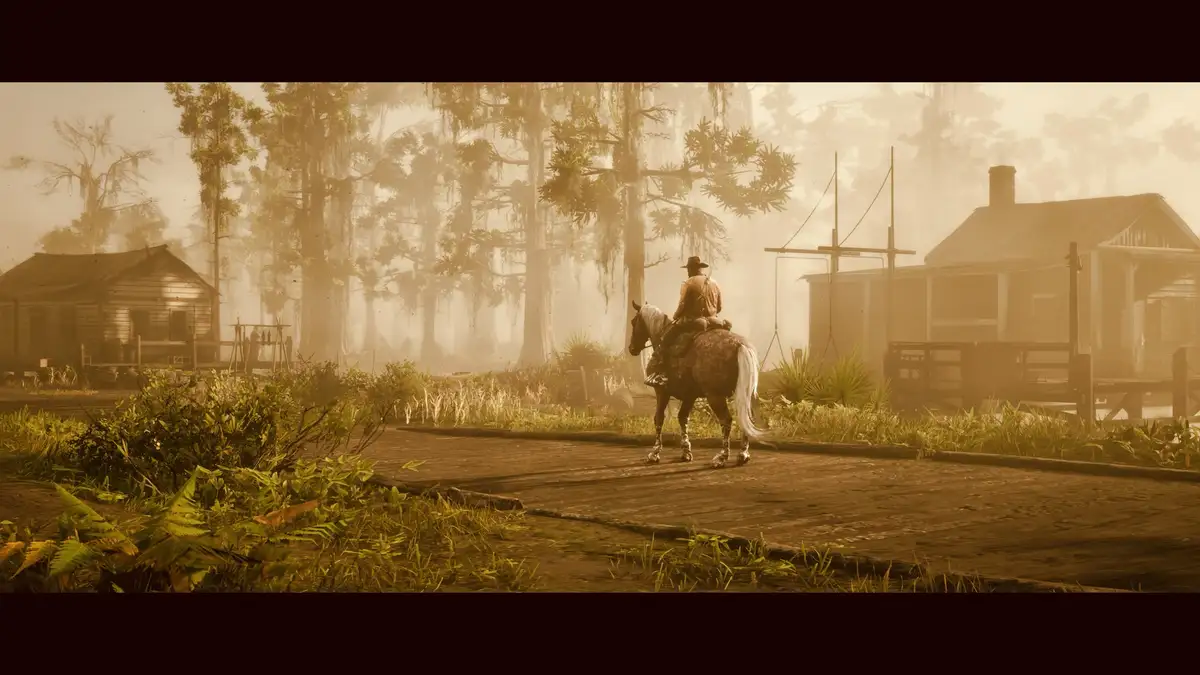 A screenshot from a video game shows a lone cowboy riding a horse down a misty, swampy road lined with wooden houses and…