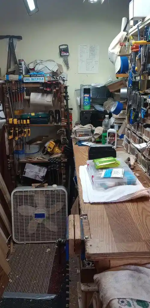 A cluttered, messy workshop filled with tools, shelves, and workbenches. A large fan sits on the floor, and various items…