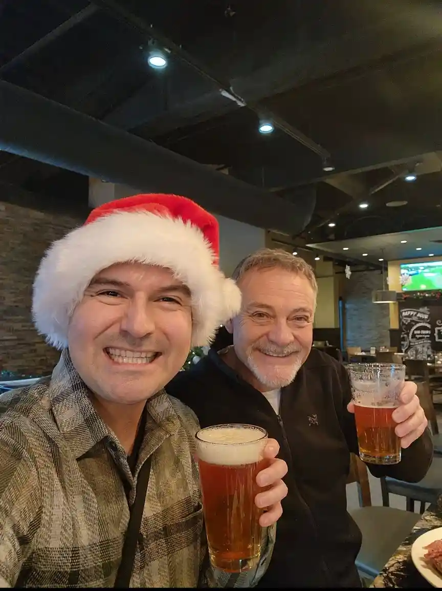 Two smiling men are posing for a selfie in a restaurant, both holding glasses of beer. The man on the left wears a red and…