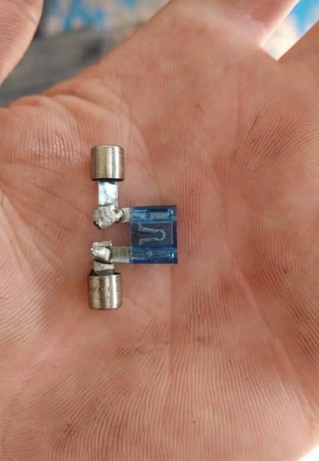 A close-up photo shows a small, broken automotive fuse held in an open palm. The fuse has a blue plastic body with a visible…
