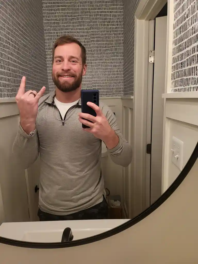 A man with a beard and a ring on his finger takes a selfie in a bathroom mirror while making the 'rock on' hand gesture with…