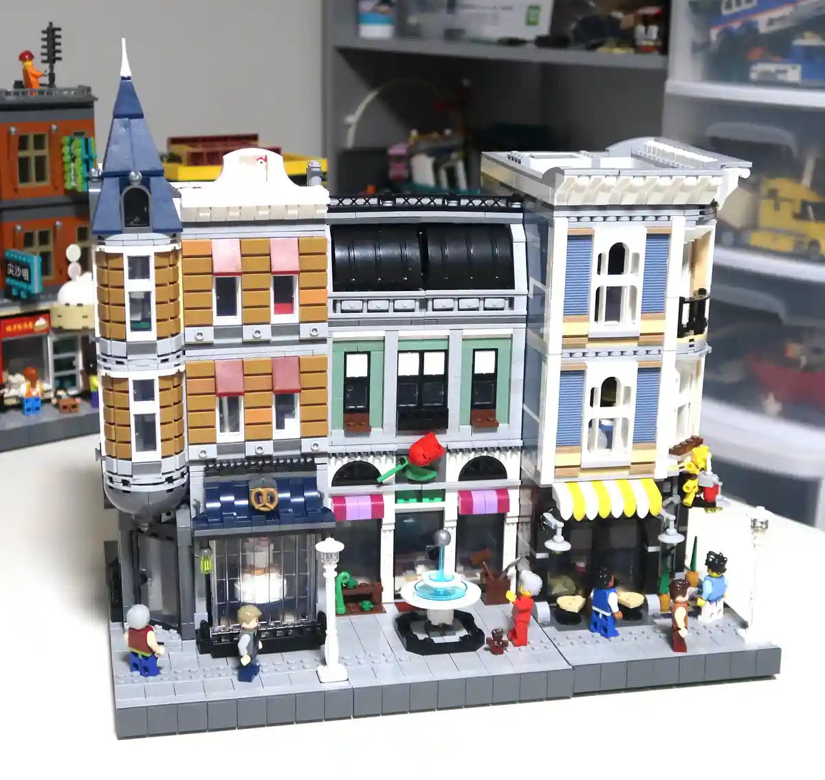 A detailed Lego city street scene with multiple buildings, tiny Lego figures, and a central fountain, overlaid with the text…