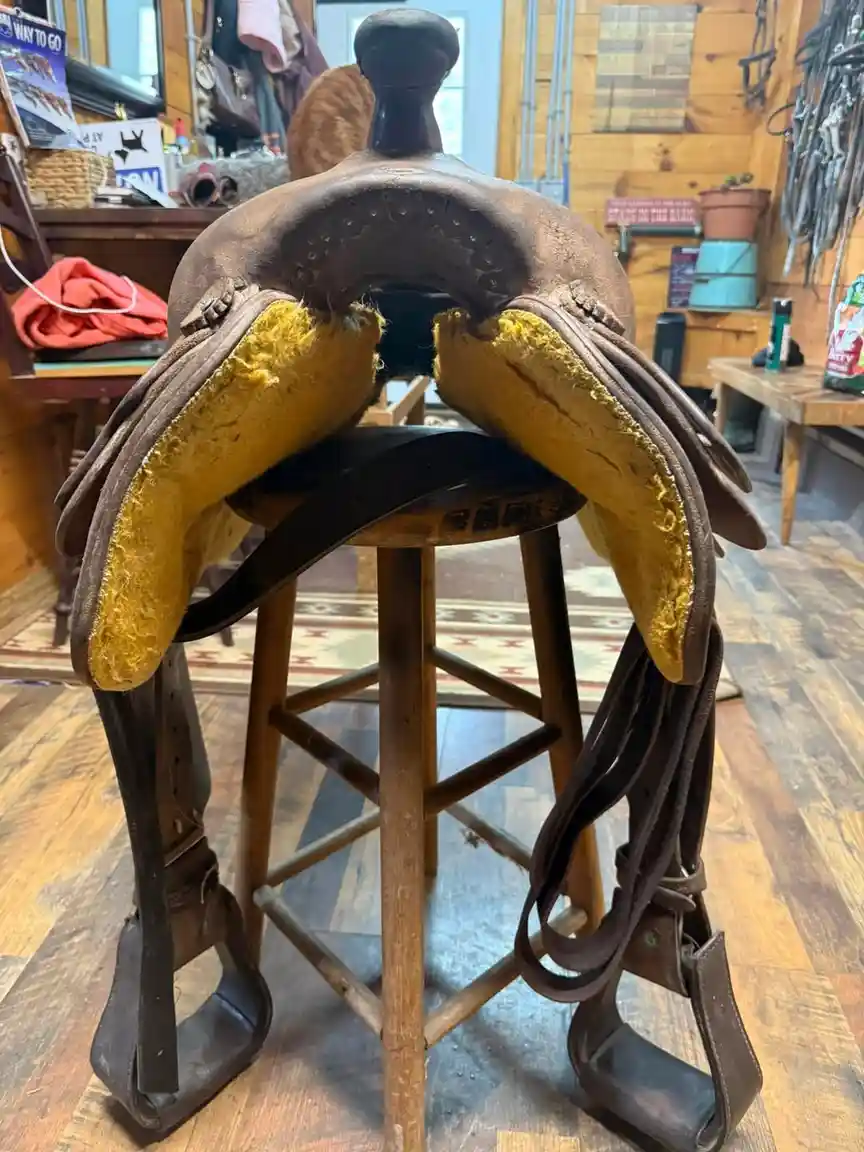 A well-worn brown leather horse saddle with yellow foam padding is sitting upright on a wooden stool in a rustic room. The…