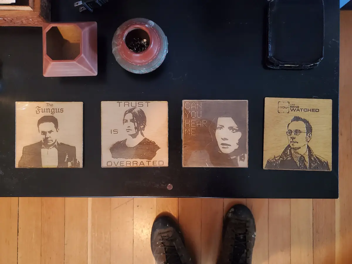An overhead shot shows four wooden coasters with engraved images and text on a black table, each depicting a character from…