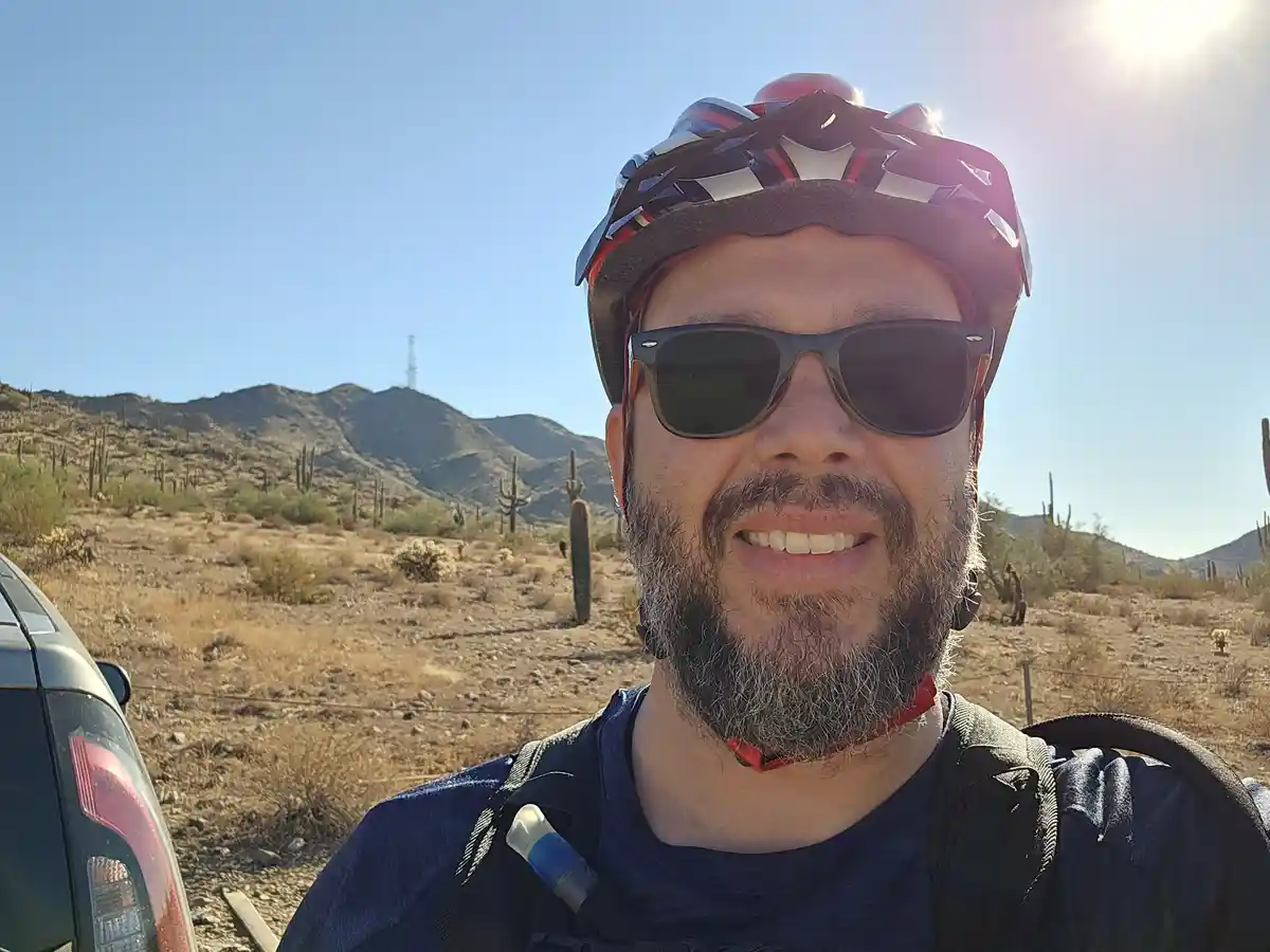 A man with a beard, wearing a red and black bicycle helmet and sunglasses, smiles at the camera while standing in a desert…