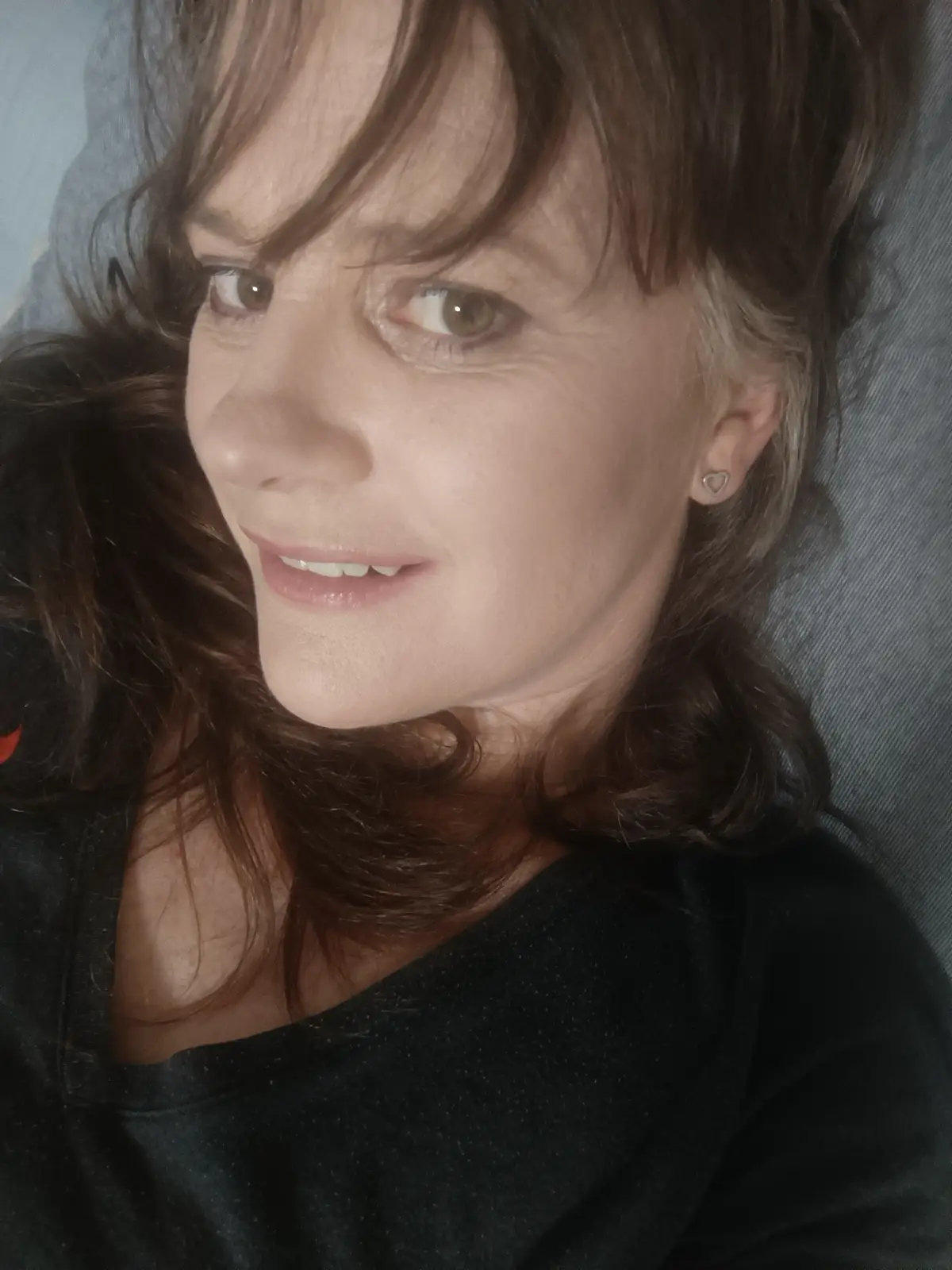 A close-up selfie of a woman with brown hair and a heart-shaped earring, smiling slightly while looking off-camera. She…