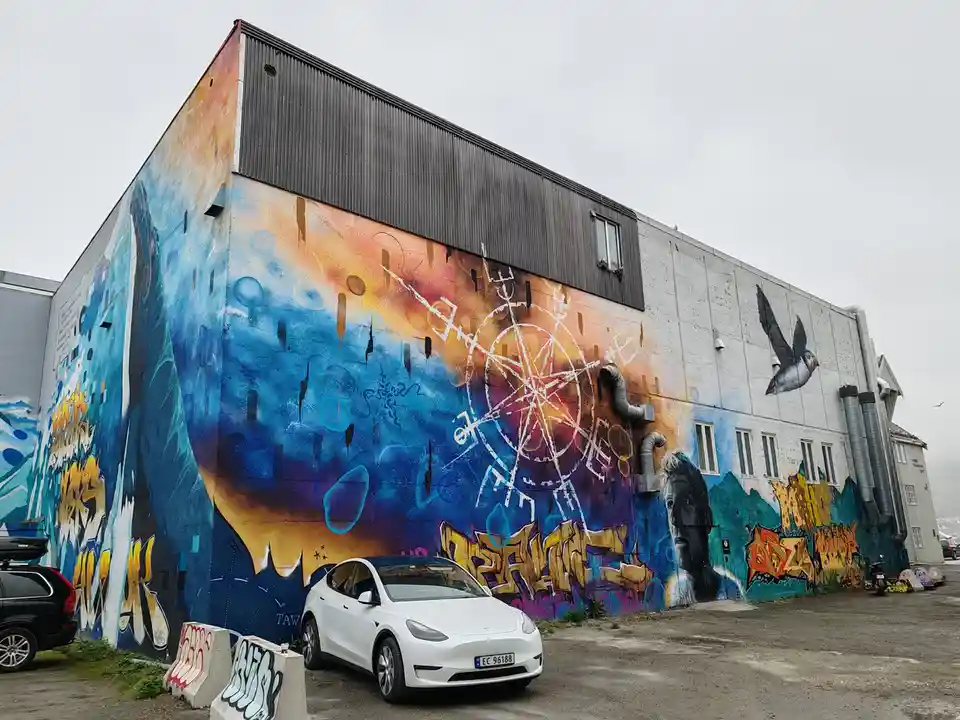 A white Tesla Model Y is parked in front of a large, colorful mural on the side of a building. The mural features abstract…