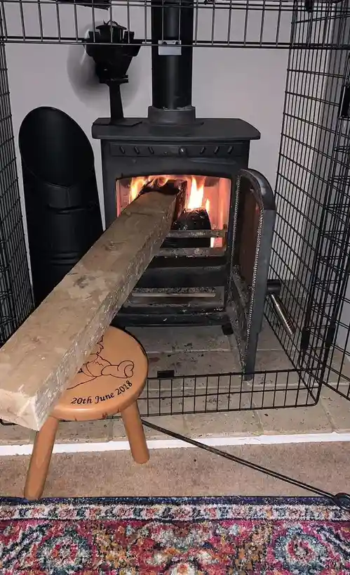 A large, unburned log is jammed horizontally into the open door of a lit wood-burning stove, with the log resting on a small…
