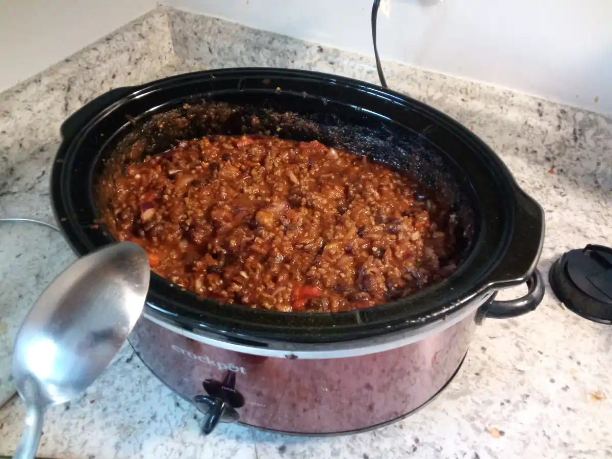 A black Crock-Pot slow cooker sits on a speckled countertop, filled with a thick, reddish-brown chili containing visible…