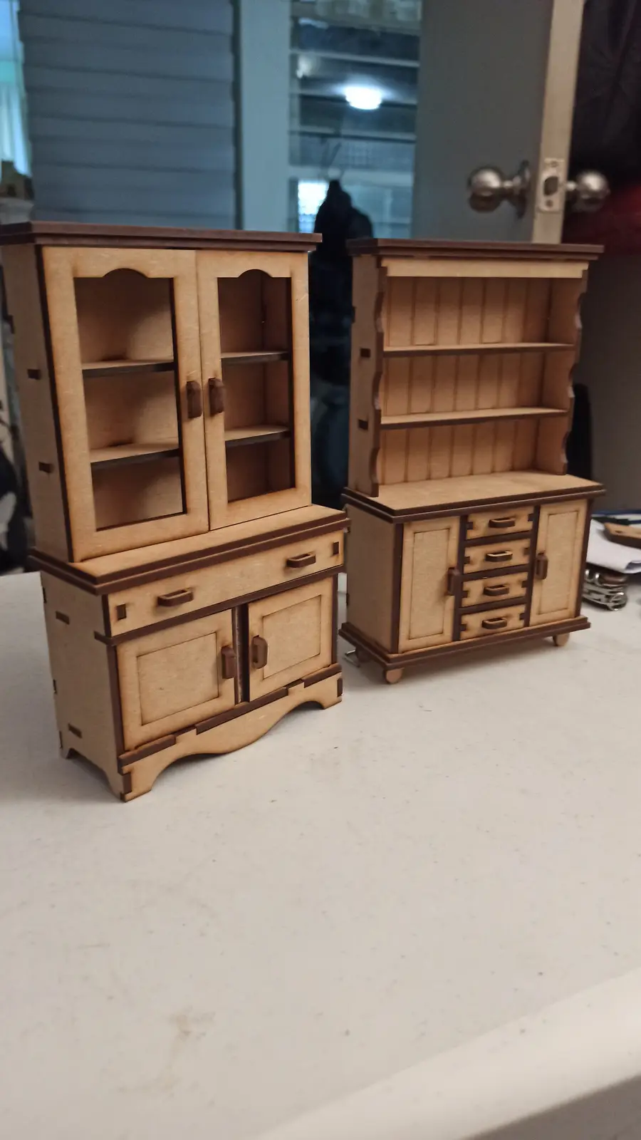 Two miniature wooden furniture pieces, one a hutch with glass doors and drawers, the other a bookcase with shelves and…