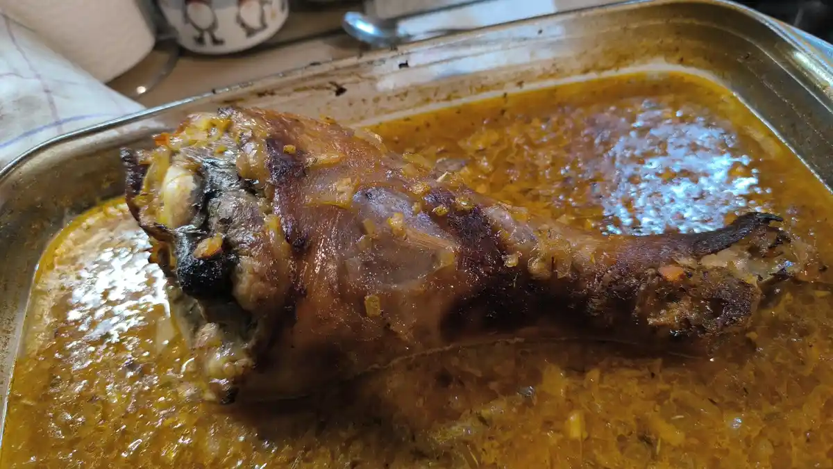A close-up photo shows a roasted chicken leg in a metal baking tray, partially submerged in a golden-brown, herb-speckled…