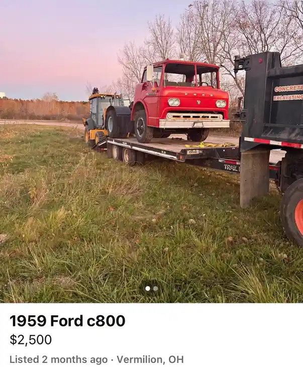A vintage red 1959 Ford C800 truck is being transported on a flatbed trailer pulled by a yellow skid-steer loader, parked in…